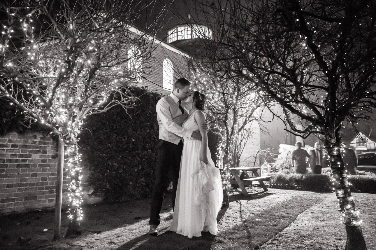 Black and white image of a wedding couple kissing amongst trees wrapped with fairy lights