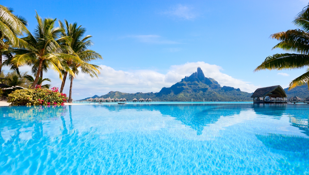 A South Pacific island rises out of the sea, with overwater bungalows and a deserted beach