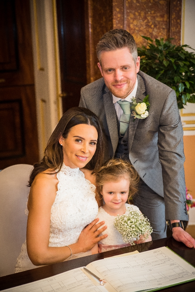 Bride, groom and daughter smile at wedding register