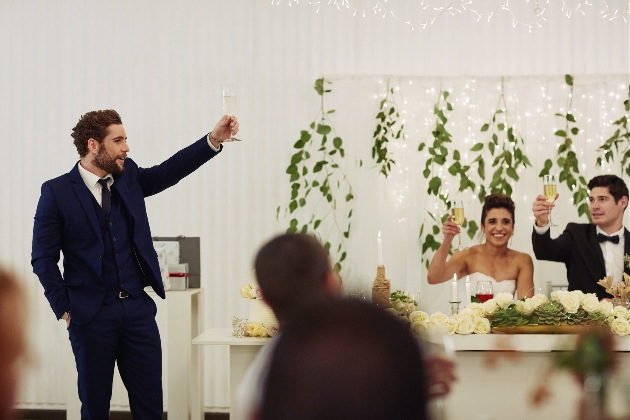 best man giving a speech at a wedding standing up while bride and groom sat down at top table