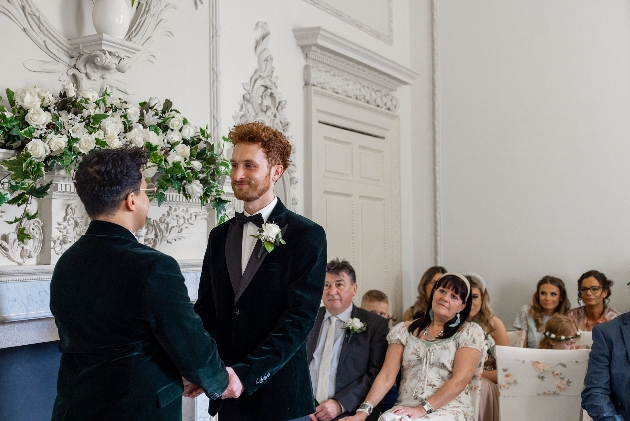 two grooms holding hands at the end of an aisle with guests watching