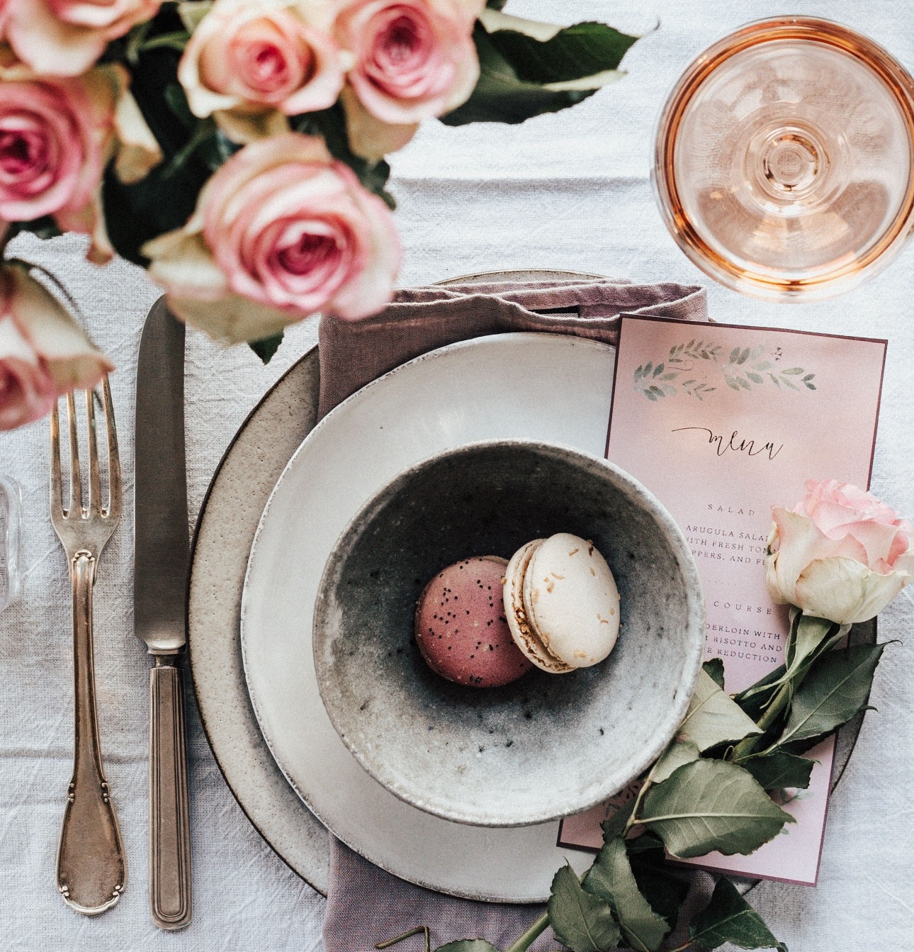 bowl of pretty macarons on charger plates with cuttlery napkin and menu, glass of wine next to it