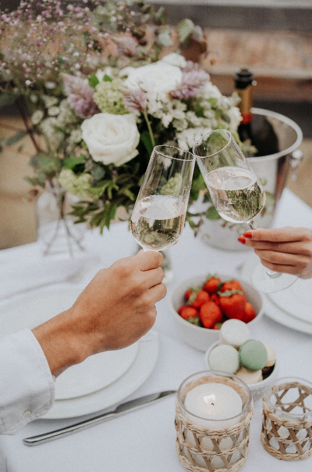 two people gentle holding to champagne glasses together at a dinner table with strawberries and macarons on
