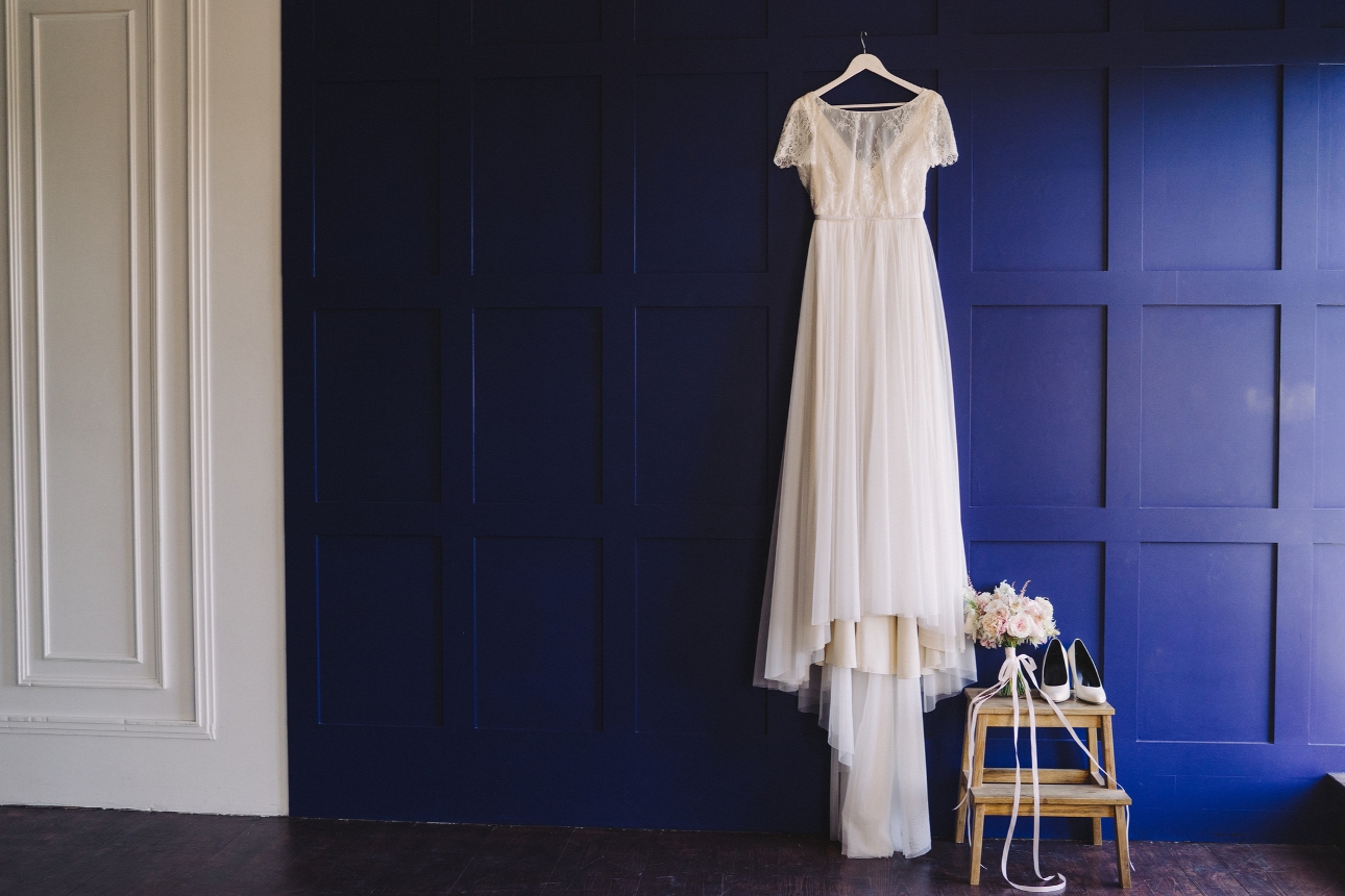 wedding dress hung on a blue panelled wall with shoes and bouquet on the floor