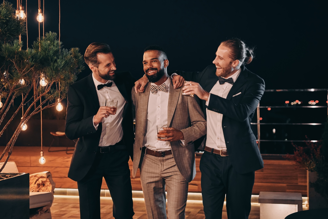 three wedding guests in suits holding drink at night on a balcony