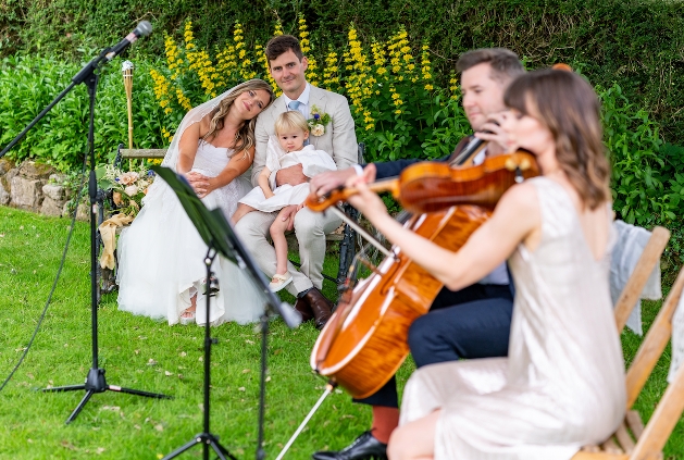 bride and groom sat down holding their child on their laps watching the string quartet