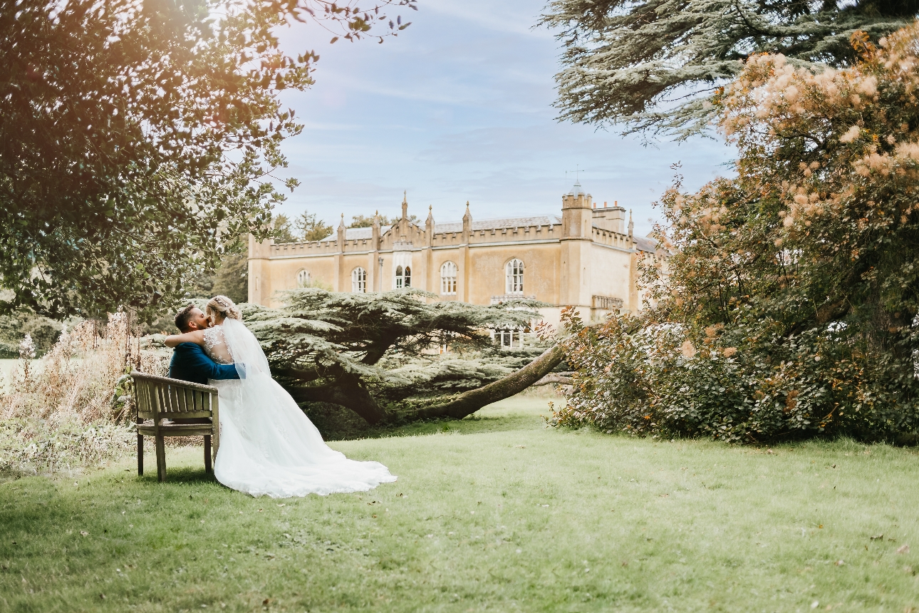 Dream wedding venue historic house in background of gardens with bride and grooms kissing on a bench