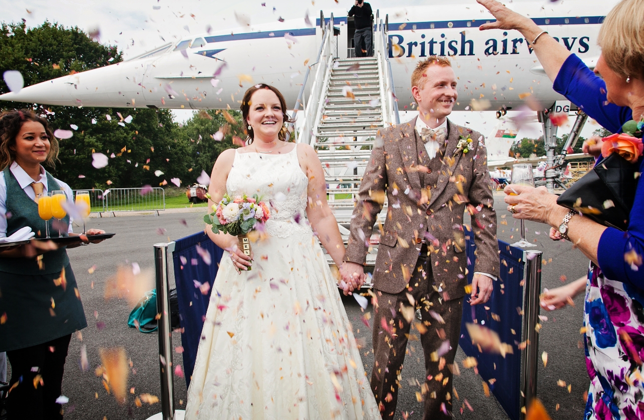 birde and groom in front of plan having their guests throw confetti on them