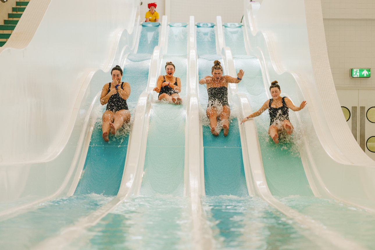 four ladies in black cosies sliding down four slides in pool