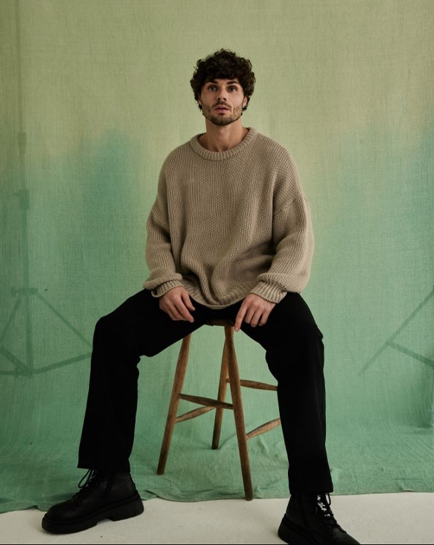 man in cream jumper and black trousers on a stool in a studio with curly hair