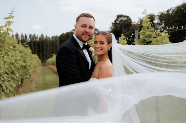A bride and groom smiling at the camera while the bride's veil blows in the wind