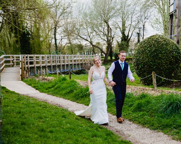 A bride and groom walking hand-in-hand along a path with a bridge in the background